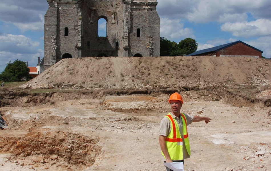 Vue du chantier de fouilles en 2010. Au premier plan un archéologue montre le cœur de l'abbatiale. En arrière plan, on peut appercevoir les 2 tours de l'abbaye du Mont Saint-Eloi.