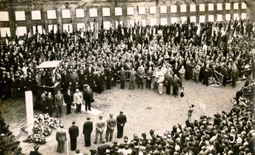 Photographie en noir et blanc d'une foule assemblée devant le poteau des fusillés fleuri pour l'occasion.