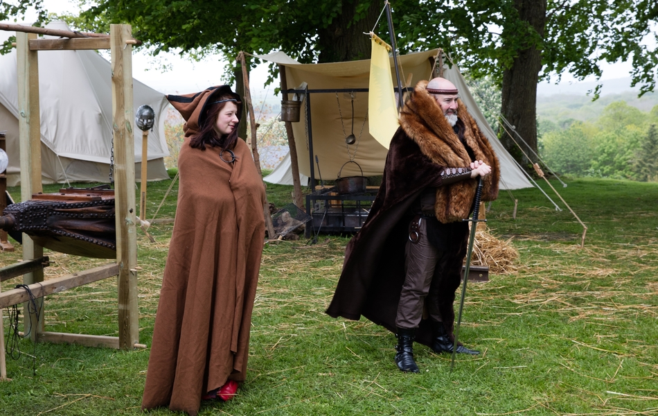 Photographie couleur montrant un homme et une femme vêtus à la mode du Moyen Age devant la reconstitution d'un campement de l'époque.