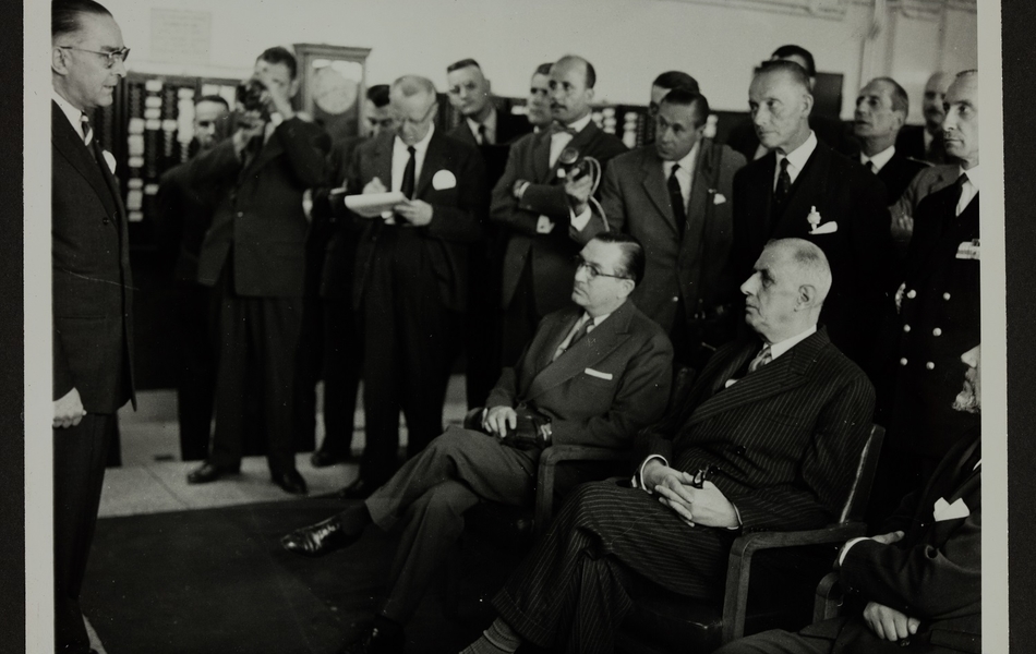 Lors de sa visite du 24 septembre 1959 à l'Usine des "Filés de Calais", le Président de la République française écoute une présentation. Des journalistes enregistrent ou prennent des notes derrière lui.