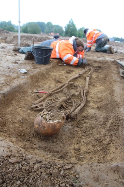 Photo de l’Archéo-anthropologue du Pas-de-Calais dégageant la terre des pieds d’un squelette. Le squelette est allongé et son crâne est endommagé.